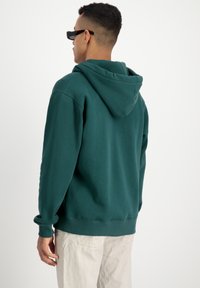 Sudadera en azul verdoso oscuro hecha de una tela suave, con capucha ajustable y puños acanalados. El modelo lleva pantalones claros.