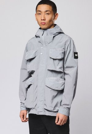 Ung mand iført en lysegrå hættejakke med flere klaplommer og en sort lap på venstre ærme, stående mod en grå baggrund.