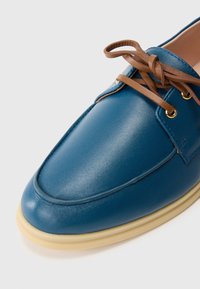 Blue leather shoe with beige sole and brown laces tied in a bow, shown in close-up on a white background.