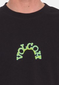 Sort bomulds t-shirt med et fremtrædende, buet gulgrønt "VOLCOM" logo på forsiden. Rund halsudskæring og korte ærmer.