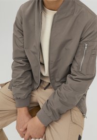 Chaqueta bomber gris con bolsillos con cremallera, puños acanalados, textura suave y un diseño sencillo, llevada sobre un suéter beige y pantalones.