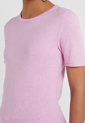 Close-up van een persoon die een effen lichtroze T-shirt met korte mouwen draagt en een enkele kleine oorring op een neutrale achtergrond.