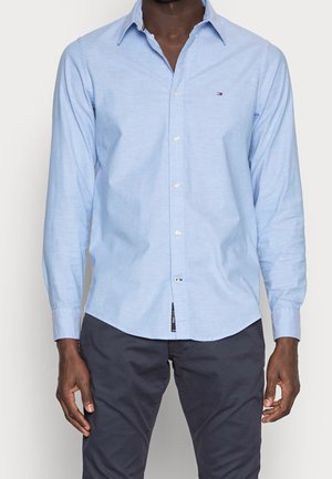 Mann trägt ein hellblaues Hemd mit Knöpfen und kleinem Logo auf der Brust sowie eine dunkelgraue Hose und steht vor einem einfarbigen Hintergrund.