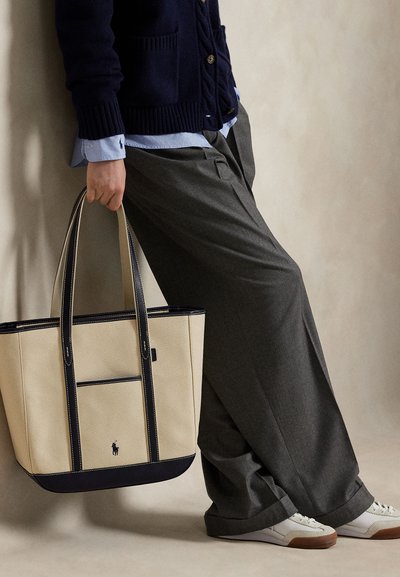 Person i grå vidbenede bukser og navy cardigan, der holder en beige og sort shoppertaske, iført hvide sneakers mod en neutral baggrund.