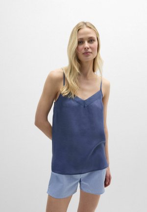 Femme blonde portant un caraco bleu sans manches et un short bleu clair à rayures, debout devant un fond clair uni.