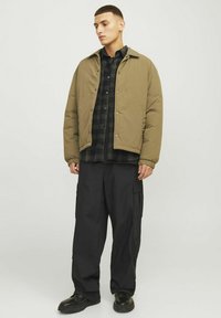 Khaki pufferjack met drukknoopsluiting, gedragen over een zwart-groen geruite blouse; gecombineerd met zwarte cargobroeken en zwarte schoenen.