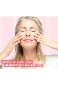 Application de produit de soin de la peau sur le visage d'une femme, avec une peau lisse, des cheveux clairs et un fond rose subtil, les mains positionnées sur les joues.