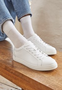 Weiße Ledersneaker mit flachen Schnürsenkeln, runder Zehenpartie und strukturierten Gummisohlen. Tragen Sie weiße Socken und blaue Jeans, auf einer hölzernen Oberfläche platziert.