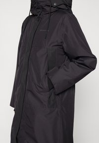 Longue veste noire en tissu imperméable, avec col montant, capuche ajustable, poches zippées et coupe d écontractée.