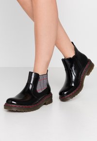 Svarta lackankelstövlar med elastiska sidopaneler i grått rutmönster, röd sömnad på sulan och strukturerad gummisula.