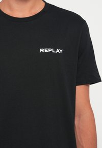 Melna kokvilnas T-kreklu ar apaļu kakla izgriezumu. Uz kreisās krūtis atrodas balts "REPLAY" logotips. Īsas piedurknes, minimāla tekstūra.