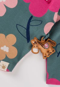 Grüner Stoff mit großen rosa und braunen Blumenmustern. Verfügt über einen orangen Pferdeanhänger aus Stoff und ein cremefarbenes Etikett mit einem braunen Spritzdesign.