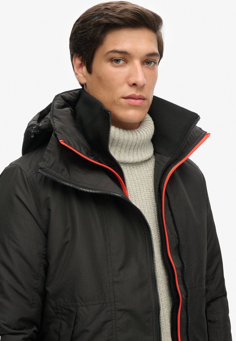 Chaqueta negra con cremallera y capucha, que presenta una cremallera naranja en contraste y una tela texturizada. Usada sobre un suéter de punto crema con cuello alto.