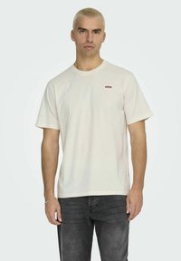 Light beige cotton t-shirt with a crew neckline and short sleeves; features a small red logo patch on the left chest and a relaxed fit.