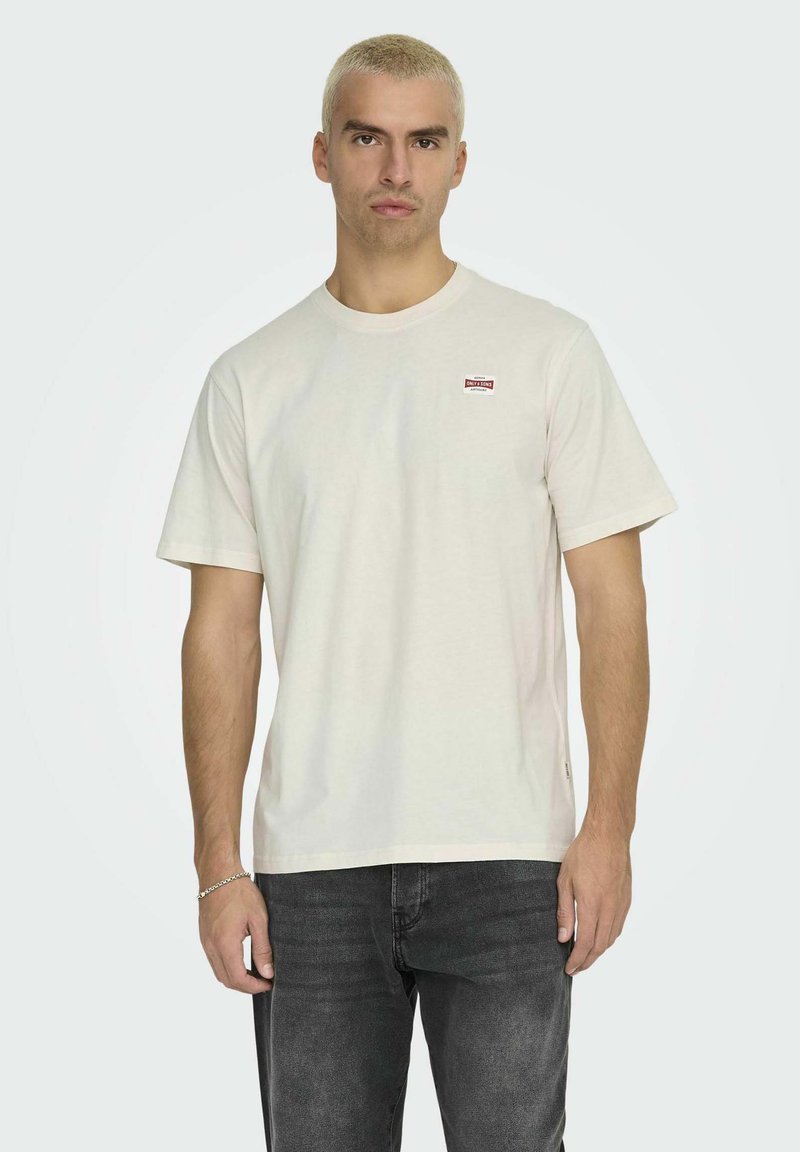 Ljus beige bomulls t-shirt med rund halsringning och korta ärmar; har en liten röd logotypficka på vänster bröst och en avslappnad passform.