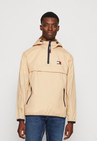 Lys beige pulloverjakke med en front halv-lynlås, hætte, marineblå nuancer og et logo-patch. Har lynlåslommer på siderne og elastiske manchetter.