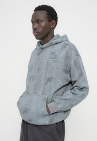 Γκρι φούτερ με διακριτικό σχέδιο από κλαδιά δέντρου, με εμπρόσθια τσέπη, κουκούλα με κορδόνια και ριμπ μανσέτες. Μαλακό, υφασμάτινο ύφασμα με υφή.