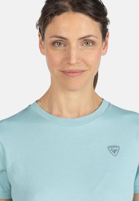 Světle modré bavlněné tričko s kulatým výstřihem, krátkými rukávy a malým šedým logem na levé hrudi. Hladká textura a neformální design.