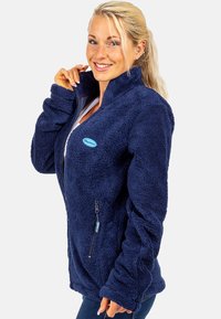 Donkerblauwe fleece jas met hoge kraag, volledige rits en zijzak. Zachte textuur en een ontspannen pasvorm, met een lichtblauw merkembleem.