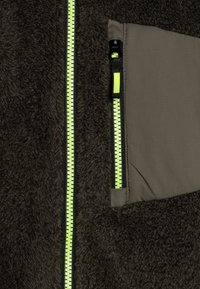 Mørkebrun fleecejakke-seksjon med en fremtredende neongrønn glidelås, ved siden av en glatt stofflapp med en liten lomme og glidelås.
