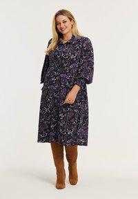 Lächelnde Frau, die ein schwarzes, blumiges Midi-Kleid mit Puffärmeln und braunen knielangen Wildlederstiefeln trägt, steht vor einem einfarbigen Hintergrund.