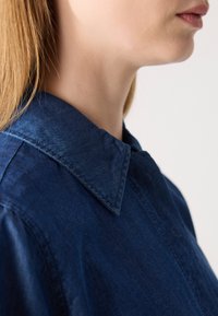 Mørkeblå denimskjorte med klassisk krage, strukturert overflate og synlige sømmer langs kragen og knappeåpningen.