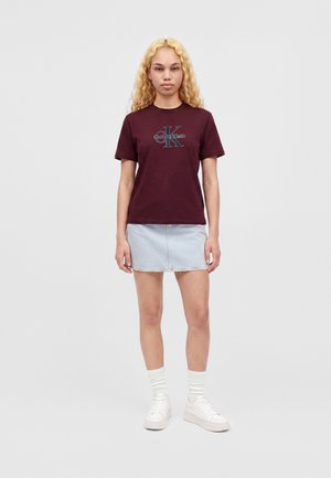 Jeune personne aux longs cheveux blonds bouclés, portant un T-shirt Calvin Klein rouge foncé, une jupe en jean claire, des chaussettes blanches et des baskets blanches, debout immobile.