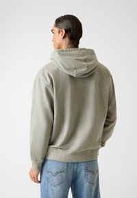 Mikina s kapucňou z mäkkej zelenej tkaniny, s voľným strihom a rebrovanými manžetami. Kombinovaná so svetlomodrými džínsami s dekoratívnym prešívaním.