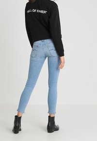 Mujer con jeans ajustados de color azul claro, botas negras hasta el tobillo y un sudadera negra con el texto "HALL OF FAMER" en la parte de atrás, de pie de espaldas.