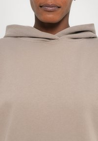 Hoodie beige clair en tissu doux et texturé avec une capuche arrondie. Le vêtement présente un design propre et simple sans logos visibles.