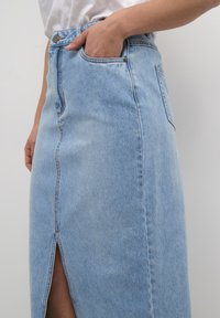 Hellblauer Jeansrock mit hoher Taille, vorderem Schlitz und Seitentasche. Der Stoff ist leicht verblasst und zeigt sichtbare Nähte.