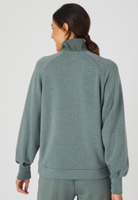 Maglione grigio-verde con colletto alto, maniche raglan e polsini a coste. Tra le caratteristiche più note ci sono la morbidezza del tessuto e la vestibilità rilassata.