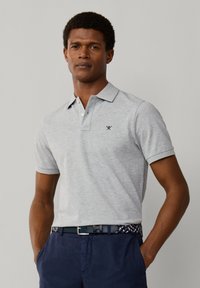Uomo che indossa una polo a maniche corte grigio chiaro e pantaloni blu navy con una cintura intrecciata, in piedi con le mani nelle tasche davanti a uno sfondo semplice.