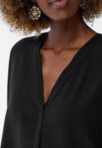 Czarna bluzka z dekoltem w serek, wykonana z lekkiego materiału; posiada subtelną teksturę i luźny krój, zestawiona z ozdobnymi, okrągłymi kolczykami w srebrnym odcieniu.