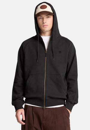 Joven con sudadera negra con capucha y cremallera, camiseta blanca debajo, pantalones marrones y gorra de béisbol beige y marrón.