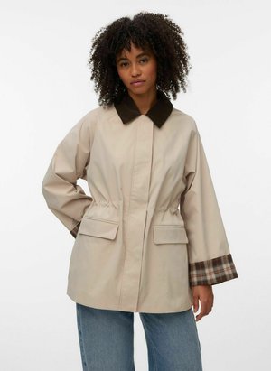 Kobieta o kręconych włosach, ubrana w beżową kurtkę z ciemnym kołnierzem i detalami w kratę na mankietach, w połączeniu z niebieskimi dżinsami, stojąca na jednolitym tle.