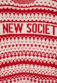 Piros és fehér kötött pulóver, ismétlődő mintával, a "NEW SOCIETY" felirattal, amely piros színben, a mellkason látható.