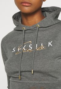 Sudadera gris hecha de tejido suave con un logo frontal en blanco y dorado, que presenta herrajes dorados en los cordones y un corte relajado.