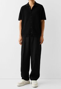 Zwarte kortemouwen shirt met een gestructureerd, wafelbreisel ontwerp en knopen aan de voorkant, gecombineerd met wijde zwarte broeken en lichte sneakers.