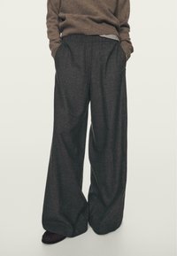 Osoba ubrana w ciemne szerokie spodnie, brązowy sweter z prążkami i brązowe buty, stoi z rękami w kieszeniach na jasnym tle.