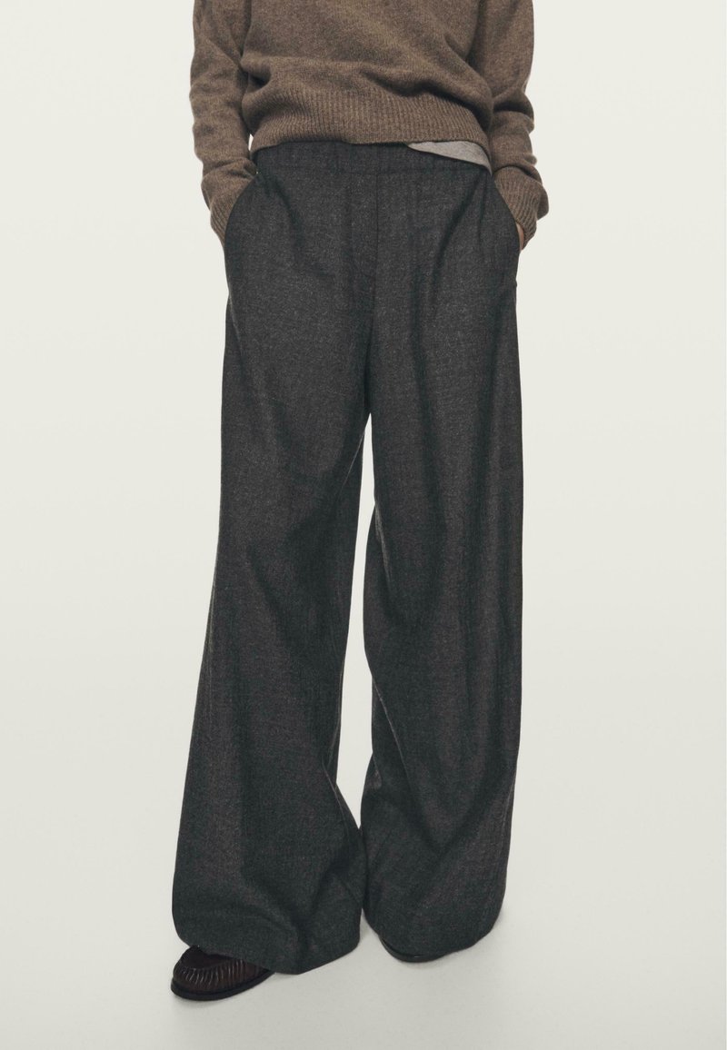 Osoba ubrana w ciemne szerokie spodnie, brązowy sweter z prążkami i brązowe buty, stoi z rękami w kieszeniach na jasnym tle.