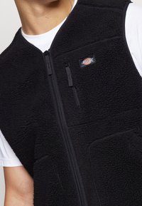 Gilet in pile nero con cerniera, caratterizzato da una piccola etichetta con logo e tessuto testurizzato. Include una tasca laterale e un design con colletto alto.