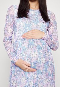 Jasnoniebieska sukienka w kwiaty z długim rękawem, z okrągłym dekoltem, elastycznym materiałem i marszczeniem podkreślającym brzuch.
