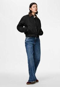 Kobieta w czarnej kurtce zapinanej na zamek z brązowym kołnierzem, niebieskich prostych dżinsach i czerwono-brązowych butach, stojąca z rękami w kieszeniach kurtki.