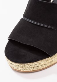 Sandale ouverte en daim noir avec une large bride et une semelle plateforme tissée en or sur fond blanc.