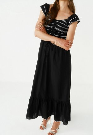 Femme en robe noire longue jusqu'aux chevilles avec des détails brodés blancs et un ourlet à volants, portant des sandales blanches à talons et des bijoux en or, bras croisés.