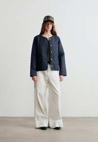 Γυναίκα που φοράει μαύρο παλτό με επένδυση, ελιάς πράσινο τοπ, λευκό φαρδύ παντελόνι, μαύρα παπούτσια και πράσινο καρό καπέλο, στέκεται μπροστά σε ένα απλό φόντο.