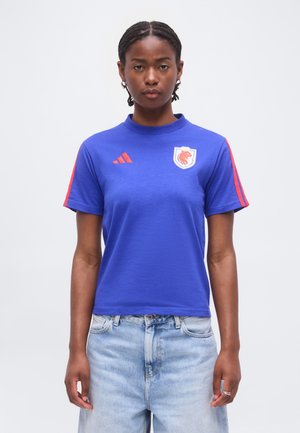 Žena s vrkočmi, ktorá má na sebe modré športové tričko s červeným logom Adidas a erbom leva, v kombinácii so svetlomodrými džínsami.