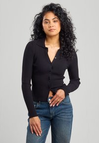 Black ribbed button-up cardigan with a collar, long sleeves, and slight crop. Worn over blue jeans, featuring a fitted design and textured fabric.