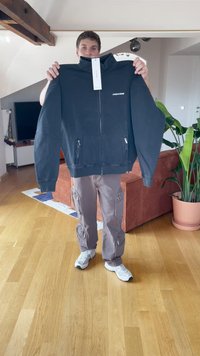 Crna zip-up dukserica s visokim ovratnikom, sadrži srebrne patentne zatvarače na džepovima i logo na grudima. U kombinaciji s sivim cargo hlačama i bijelim cipelama.